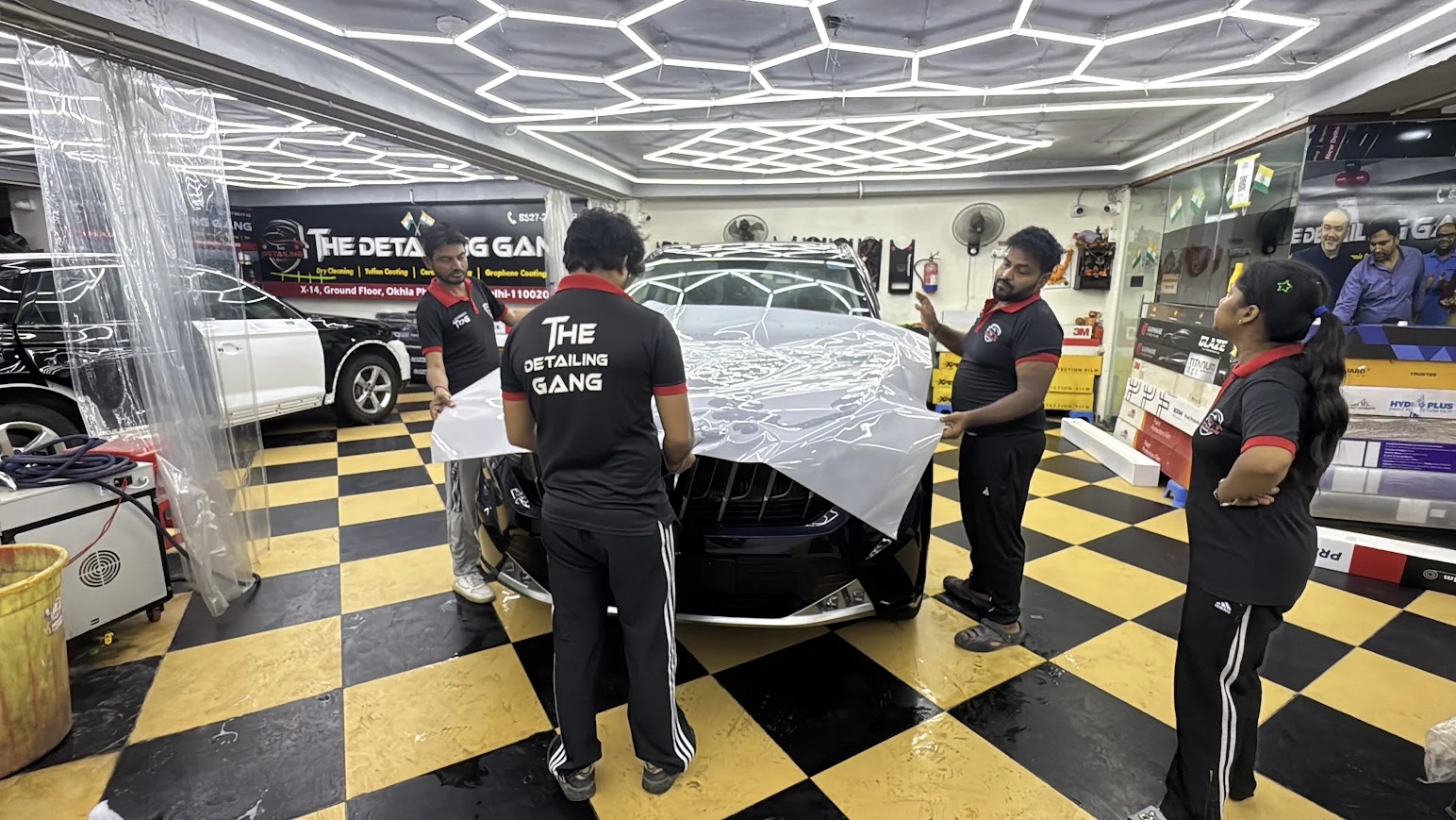 The Detailing Gang staff in black-and-red uniforms positioning PPF on a car bonnet inside the studio